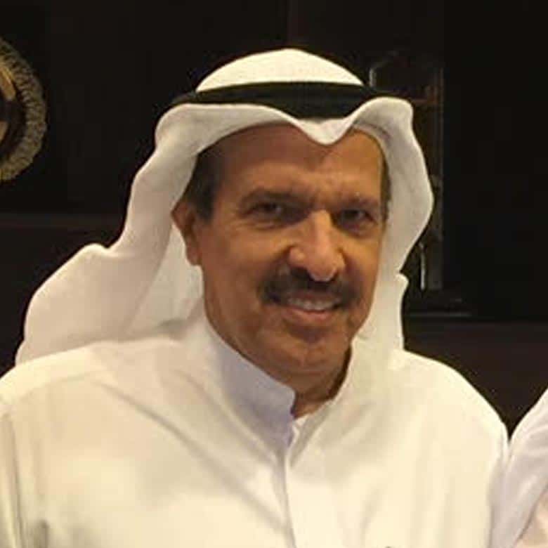 A smiling man in traditional middle eastern attire, featuring a white thobe and a black-trimmed white ghutra, exudes warmth and friendliness in an indoor setting.